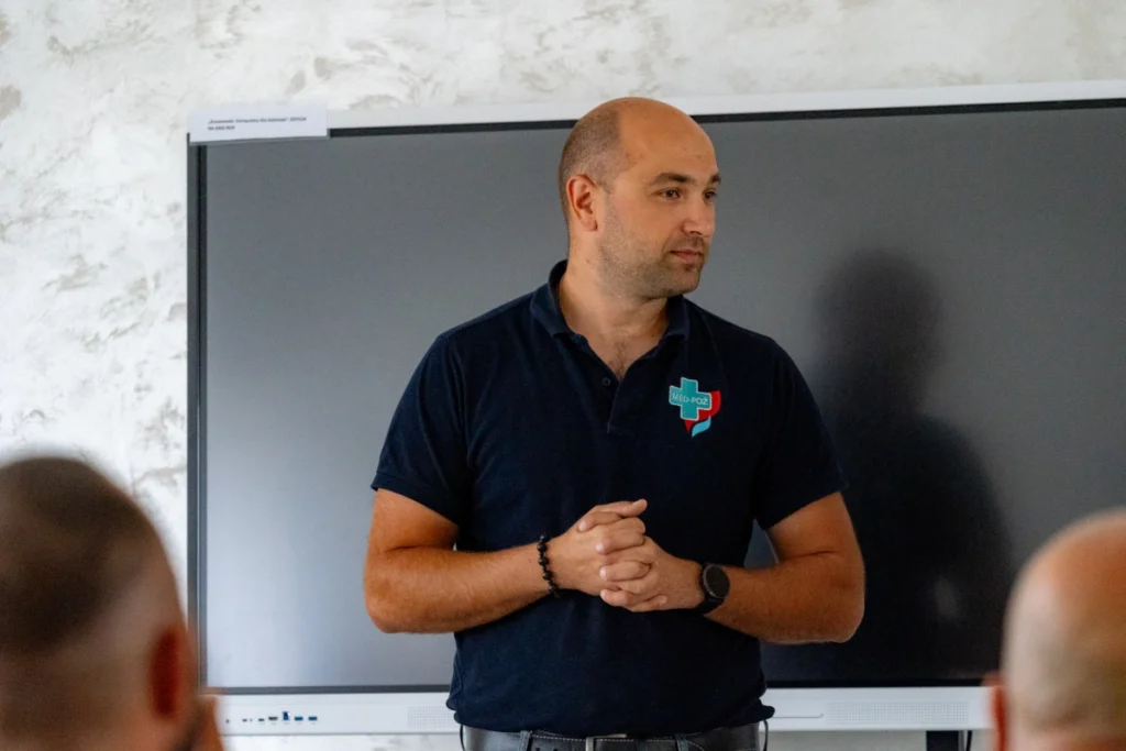 Training instructor in a navy shirt with the MED-POŻ logo stands, hands clasped, in front of the screen