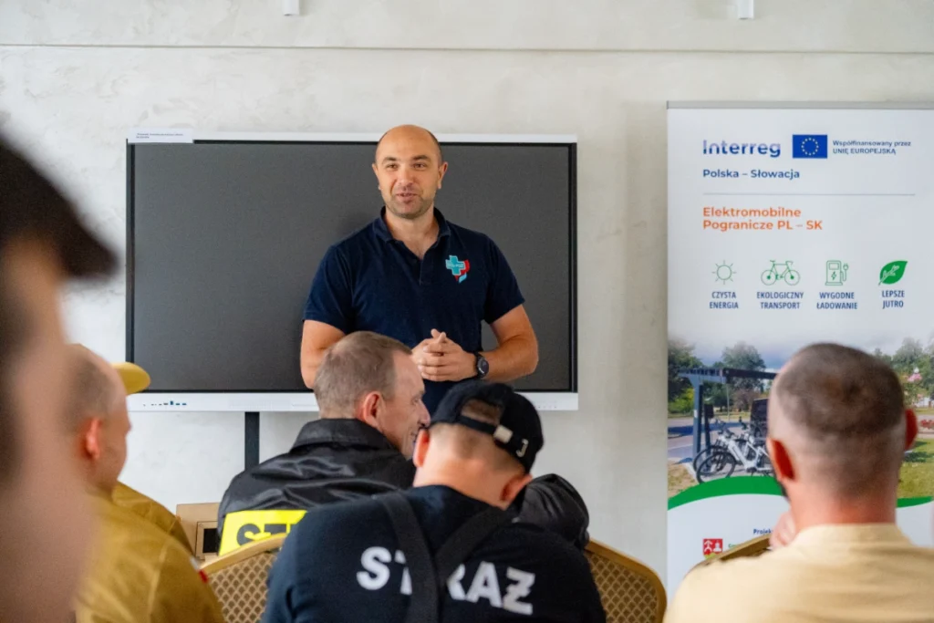 Training instructor speaks by the screen as firefighters listen, one with STRAŻ printed on his back