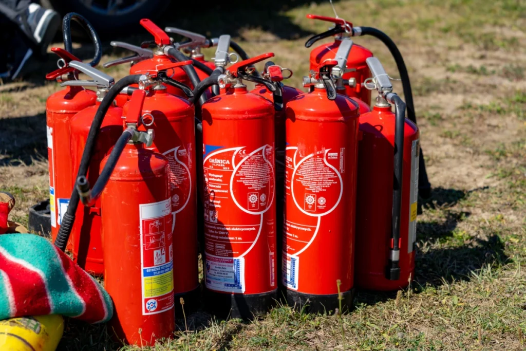 Red fire extinguishers marked GAŚNICA stand tightly grouped on the grass, a striped blanket lying beside them