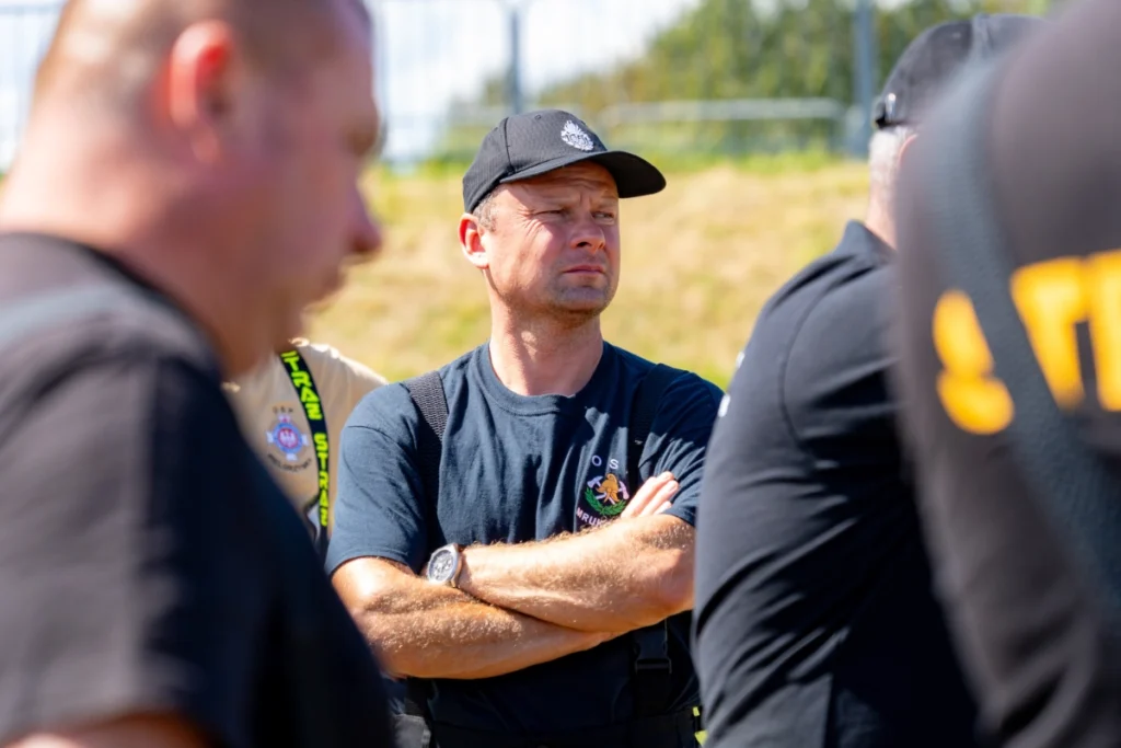 Volunteer firefighter in a navy shirt and cap listens with arms crossed, surrounded by other firefighters