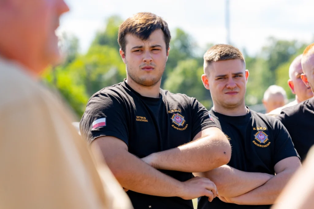 Two firefighters in black shirts reading KSRG Świerchowa listen with arms crossed