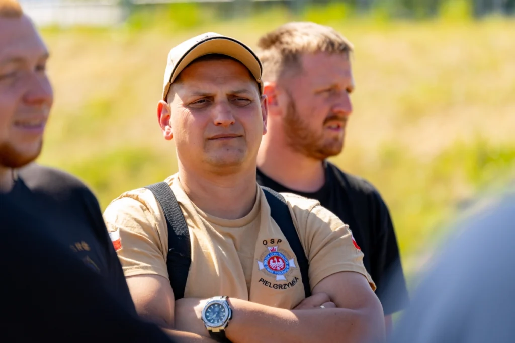 Firefighter in a sand-coloured shirt reading OSP Pielgrzymka listens with arms crossed, another firefighter behind him