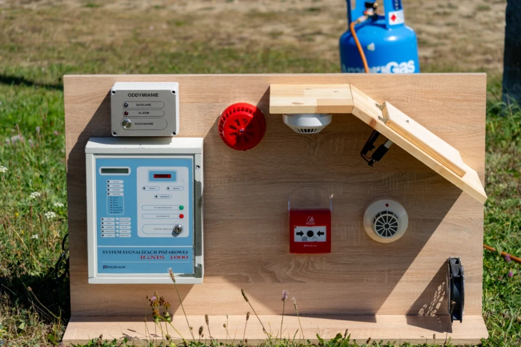 Training board on the grass with an IGNIS 1000 fire alarm control panel, detectors and a POŻAR button