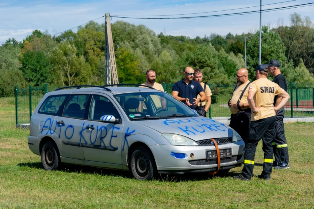 Training instructor in a navy polo shirt talks with firefighters by a silver car marked AUTO KUREK