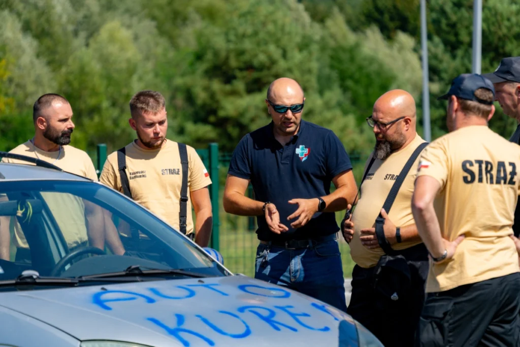 Training instructor in a navy shirt gestures, explaining to firefighters standing at the bonnet of a silver car