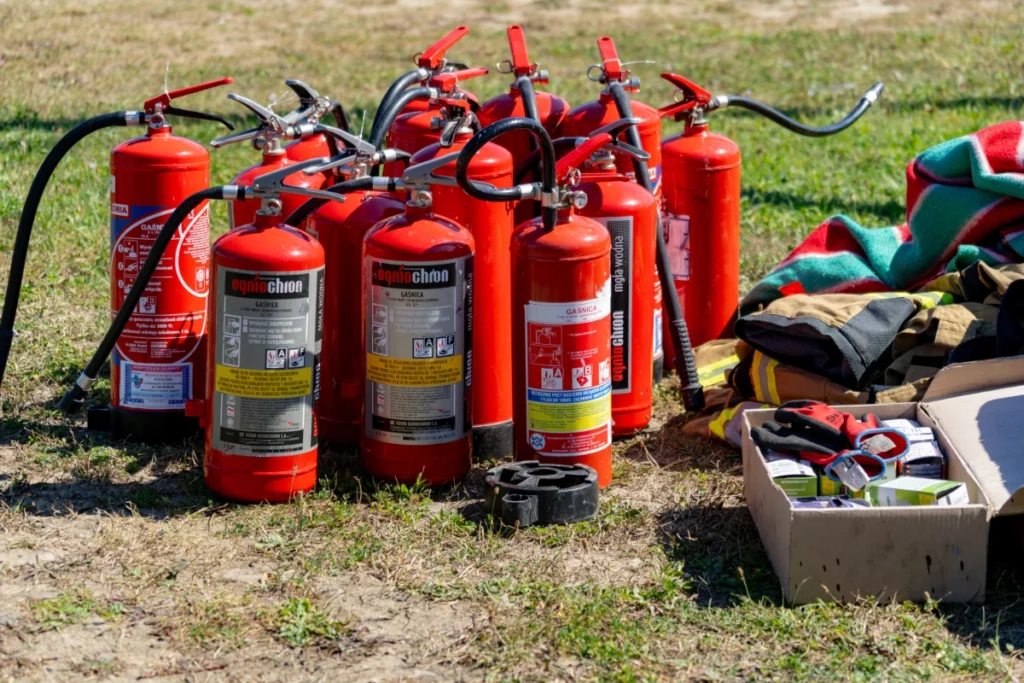 Over a dozen red fire extinguishers on the grass beside a box of gloves and folded firefighting gear