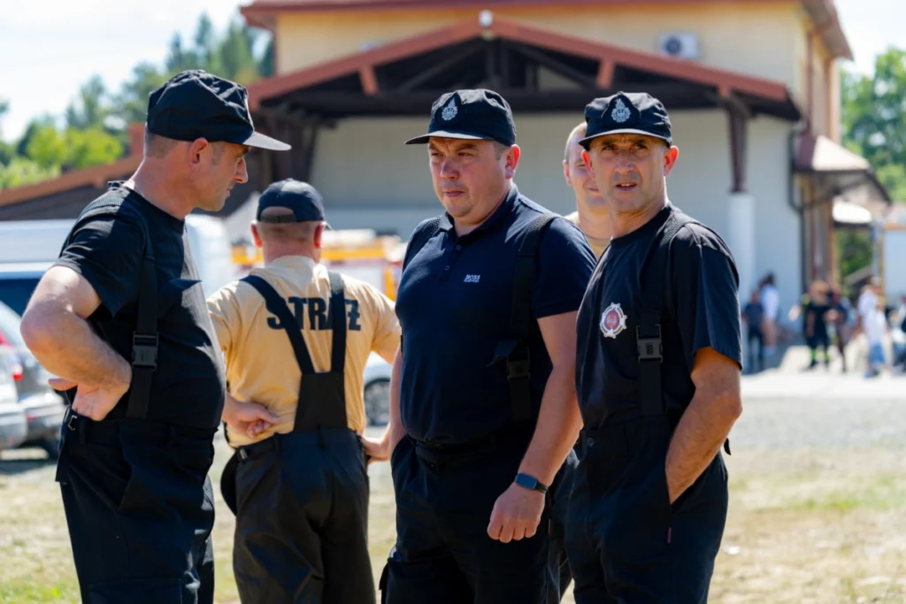 Volunteer firefighters in black uniforms talk in a group outside a building with a wooden canopy