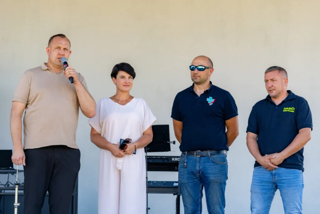 Speaker holds a microphone to his mouth beside a woman and two men, one in a HASIČI shirt