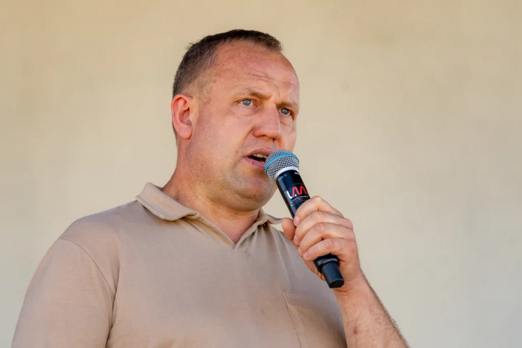 Man in a beige polo shirt speaks into a microphone on a covered stage