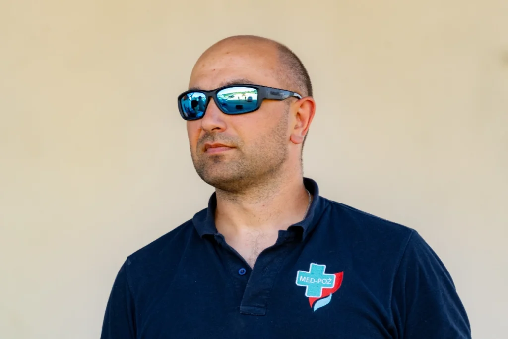 Man in a navy polo shirt with the MED-POŻ logo stands on stage listening to the speeches