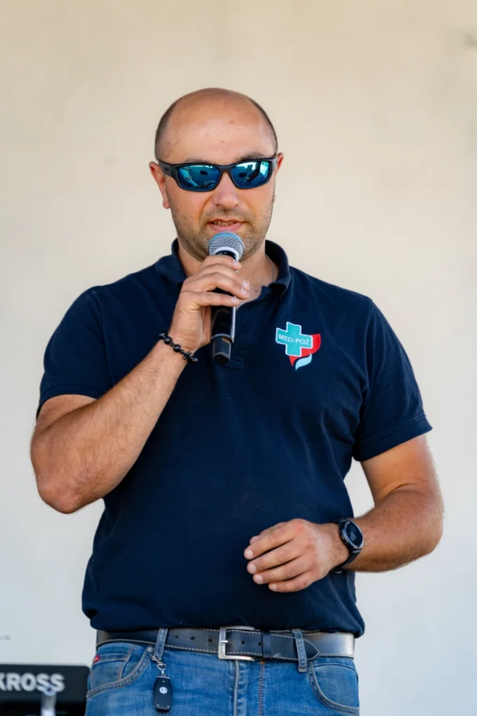 Man in a navy shirt with the MED-POŻ logo speaks into a microphone against the covered stage wall