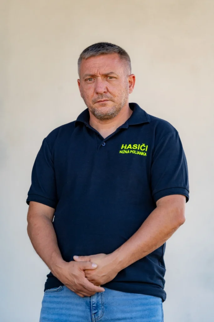 Firefighter in a navy shirt reading HASIČI NIŽNÁ POLIANKA stands with hands clasped, looking straight ahead