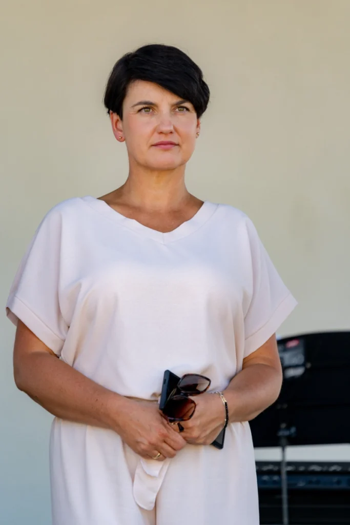 Woman in a light-coloured outfit holds a phone and sunglasses, standing on stage by music equipment