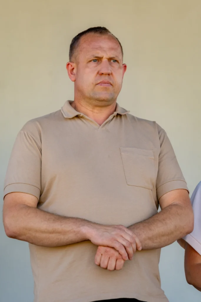 Man in a beige polo shirt stands on stage with arms folded, listening to the speeches
