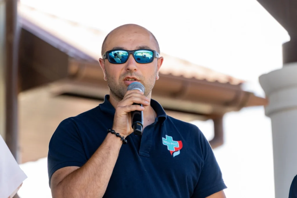 Man in a navy shirt with the MED-POŻ logo speaks into a microphone, a building's eaves behind him