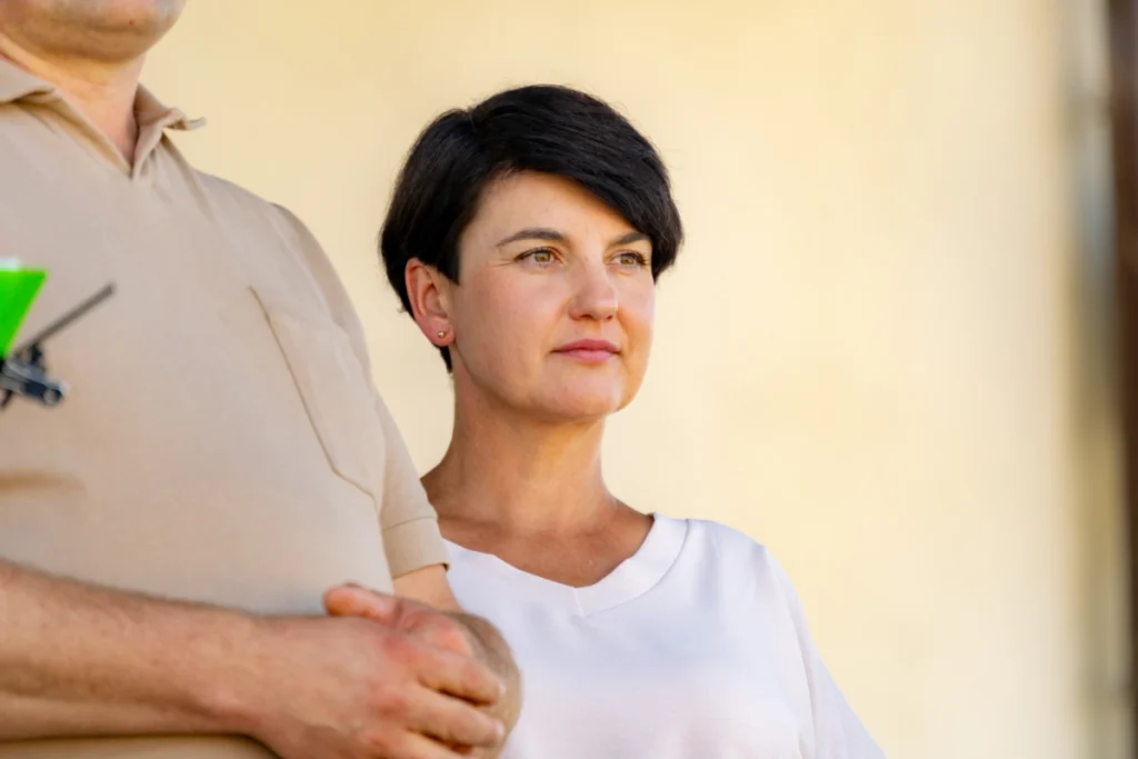 Woman in a light-coloured blouse stands on stage beside a man in a beige polo shirt, glancing sideways