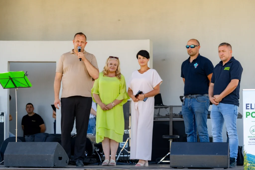 Man speaks into a microphone on a covered stage, with two women and two men standing beside him