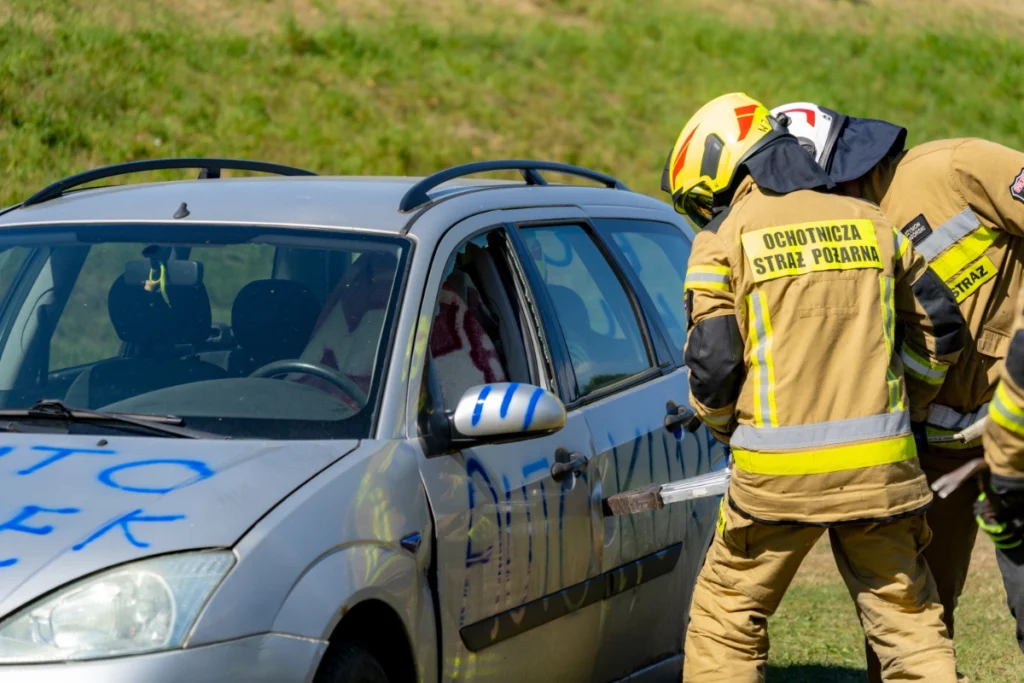Volunteer firefighter in a yellow helmet forces a silver car's front door with a tool, another firefighter nearby
