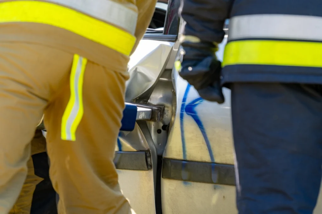 Firefighters in sand-coloured and dark uniforms wedge a hydraulic rescue tool into a car door gap, close-up