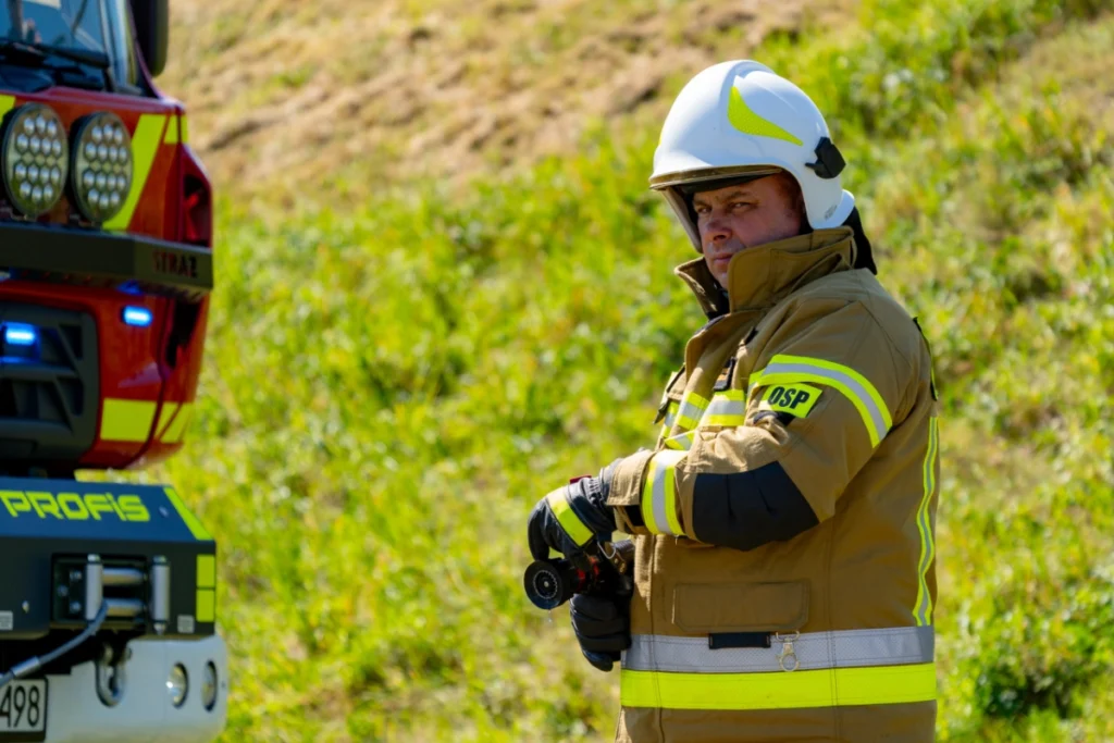 Volunteer firefighter in a white helmet holds a hose nozzle beside a fire engine, a grassy bank behind