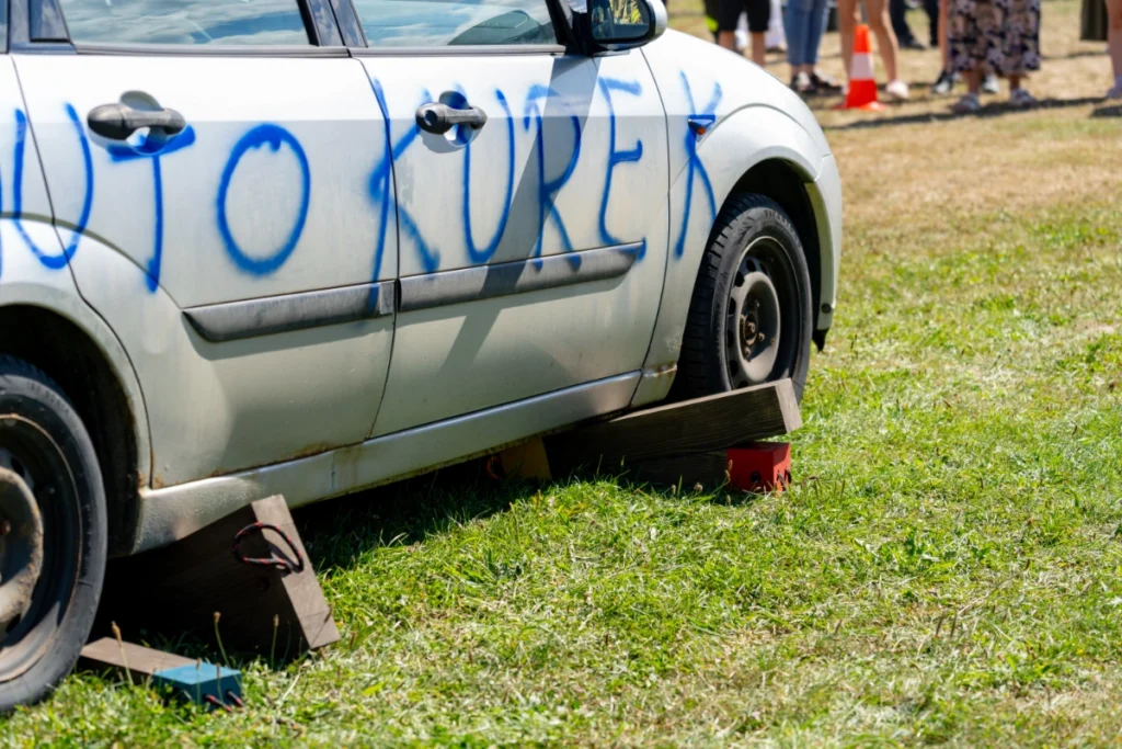 Silver car marked AUTO KUREK propped up on wooden blocks, spectators behind a traffic cone in the background