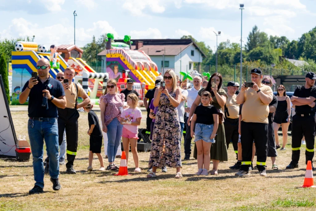Picnic visitors with children and firefighters behind cones film the demonstration on phones, bouncy castles in the background
