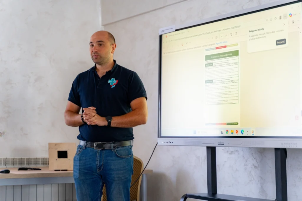 Training instructor stands by a screen showing the document “Przekazanie miejsca zdarzenia” (Scene handover), beside a wooden box