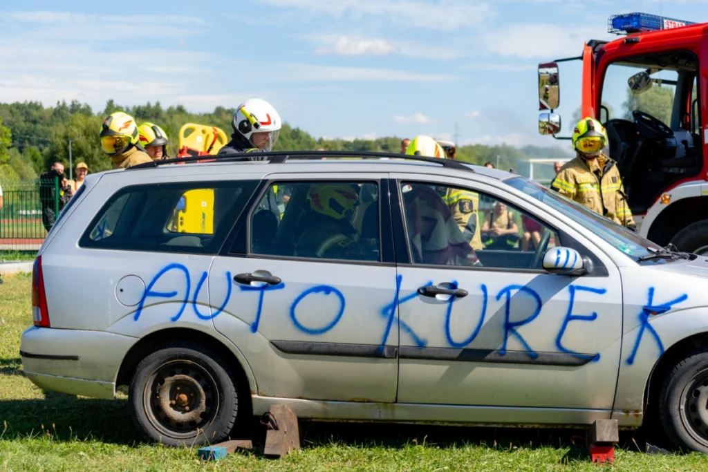 Firefighter sits in a silver car marked AUTO KUREK, behind it firefighters with a yellow spine board