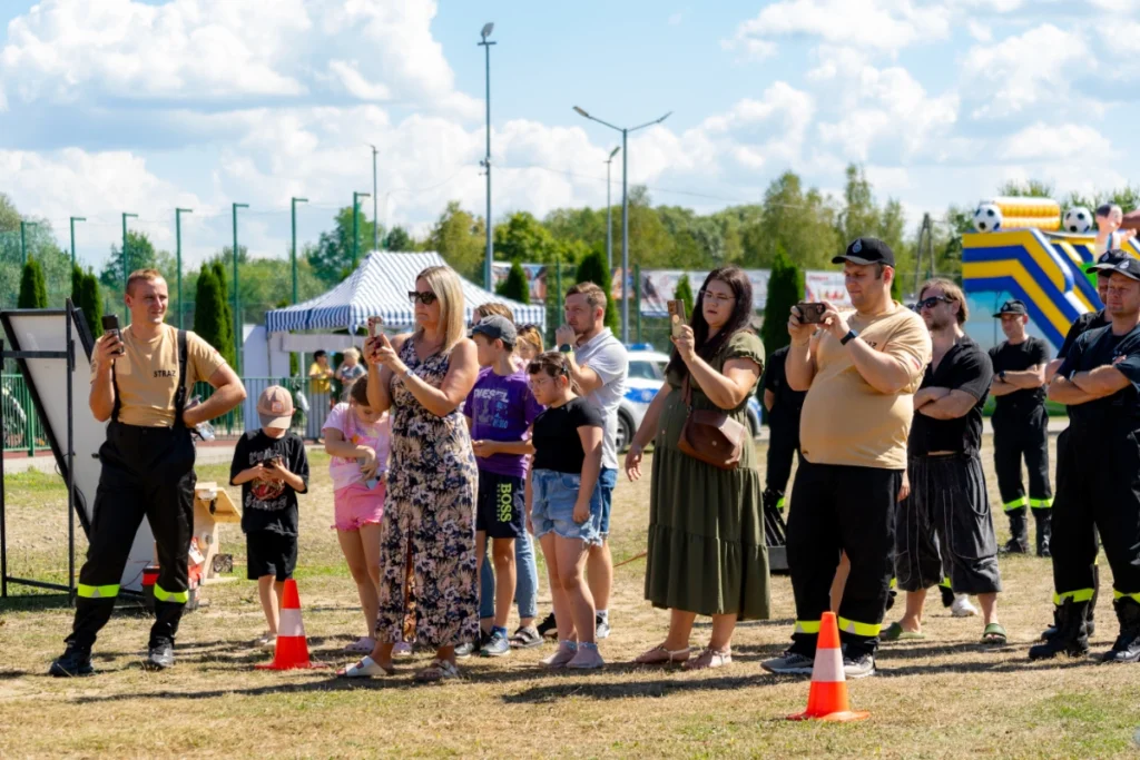 Firefighters and picnic visitors with children film the demonstration on phones, a white-and-blue tent and bouncy castle behind