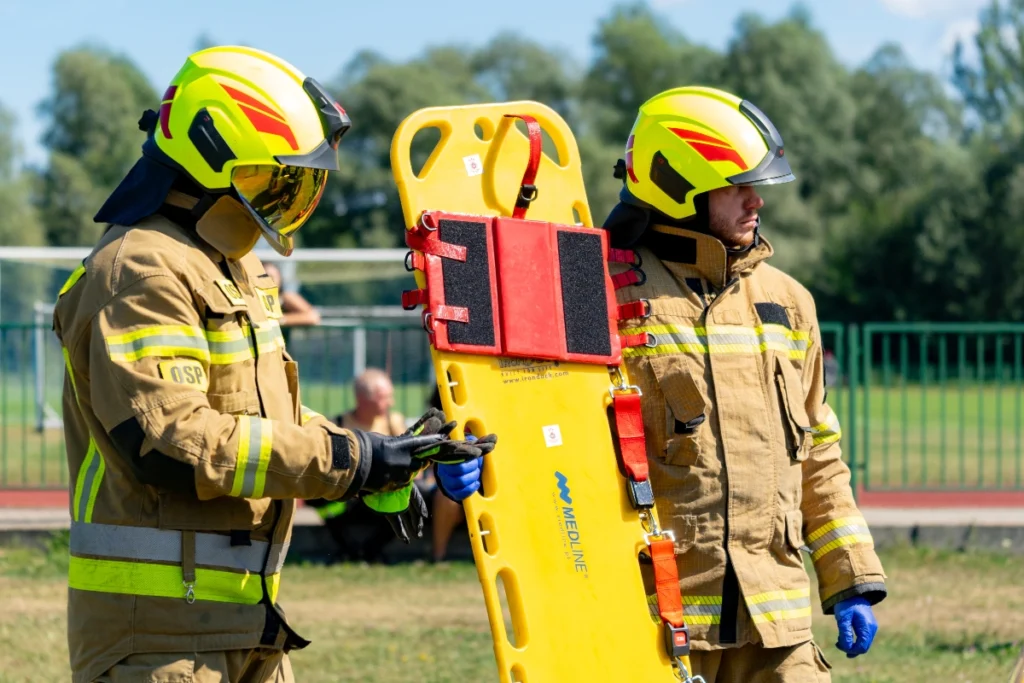 Two volunteer firefighters in yellow helmets hold upright a yellow spine board with a red head immobiliser