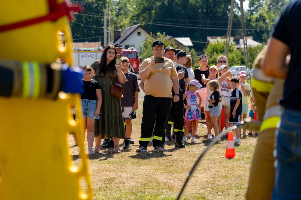 Picnic visitors and a firefighter filming on his phone watch the demonstration, yellow spine board in the foreground