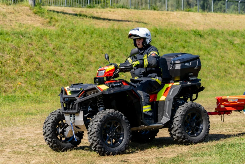 Volunteer firefighter in a white helmet rides a rescue quad with a red trailer across grassy ground