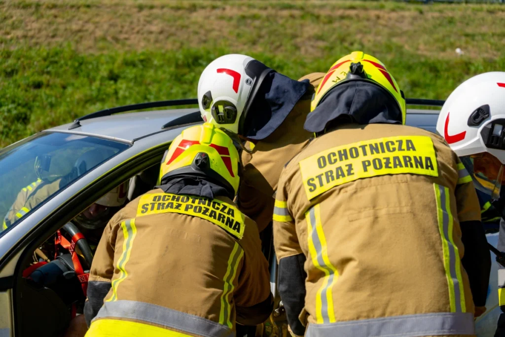 Firefighters with OCHOTNICZA STRAŻ POŻARNA printed on their backs lean over the open door of a silver car