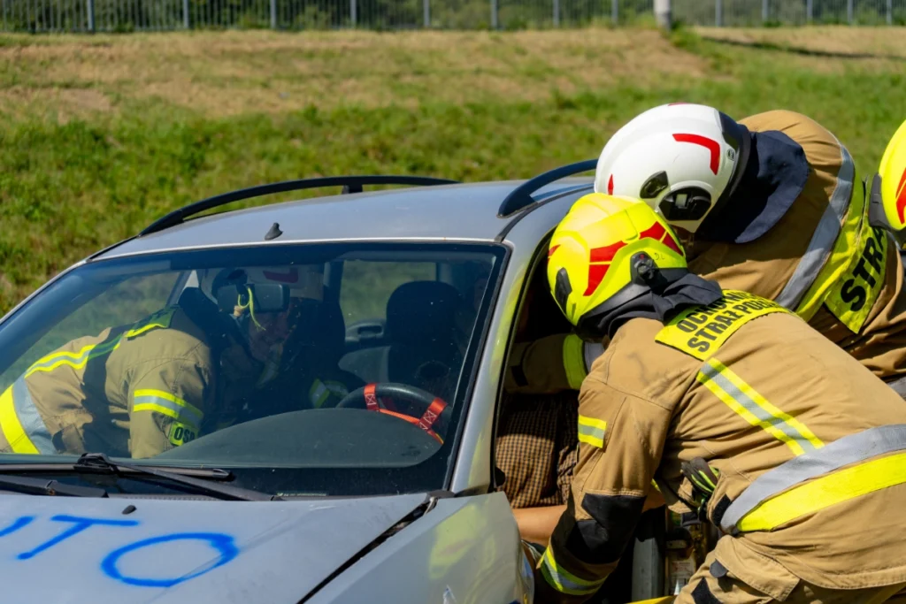 Volunteer firefighters reach through a silver car's driver door to the casualty, a firefighter sitting inside beside him