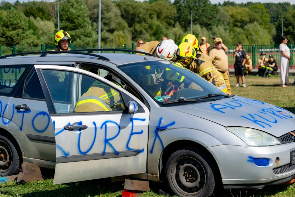 Volunteer firefighters lean over a silver car marked AUTO KUREK, with spectators standing in the background