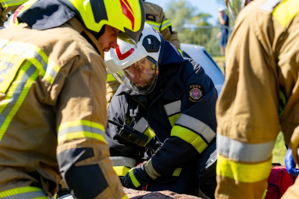 A firefighter in a white visored helmet and another in a yellow helmet bend over a casualty