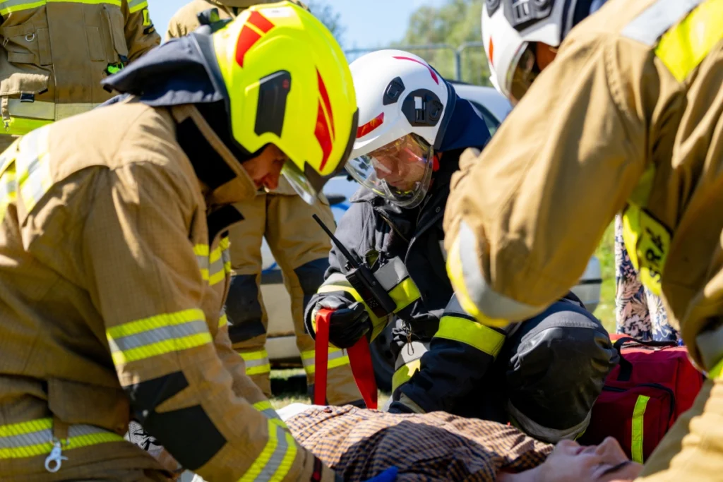 A white-helmeted firefighter holds a red strap while volunteer firefighters bend over a casualty lying on the ground