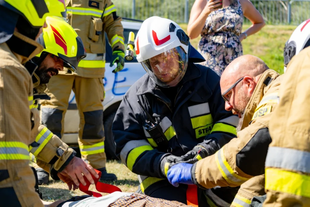 Volunteer firefighters kneel by a casualty and thread red straps, with a silver car and spectators behind