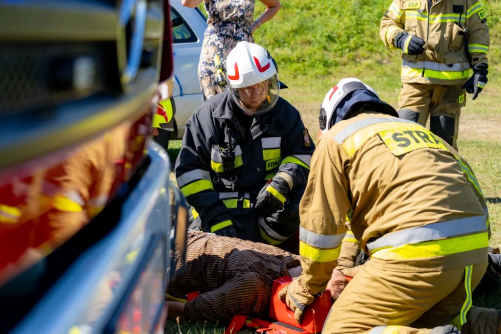 A volunteer firefighter kneels stabilising the casualty's head, next to a kneeling white-helmeted firefighter with a radio
