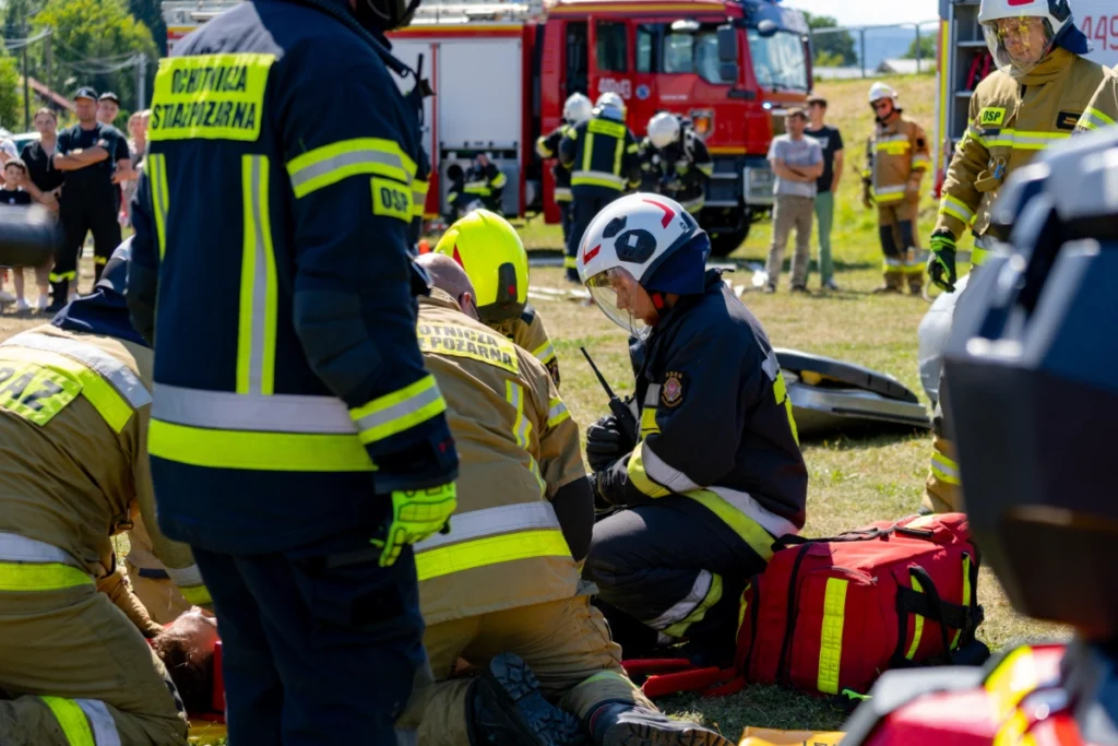 Volunteer firefighters give first aid to a casualty on the ground, red medical bag nearby, fire engine behind