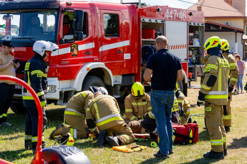 Volunteer firefighters kneel by a casualty beside a fire engine, surrounded by standing firefighters and a man watching