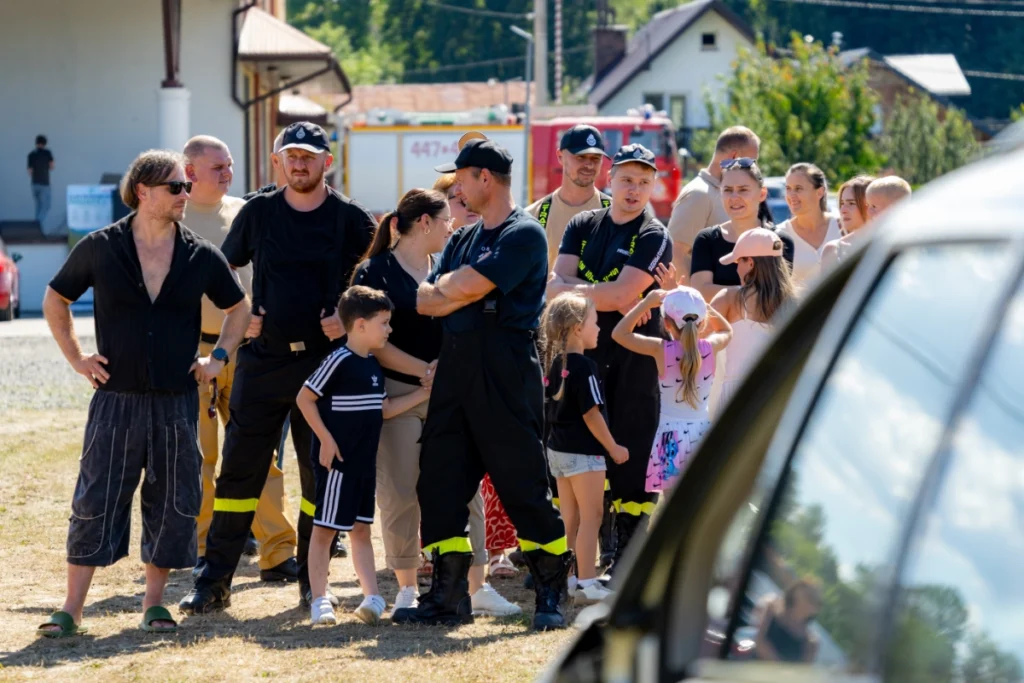 Volunteer firefighters and families with children stand on the grass watching the demonstration, with a fire engine behind