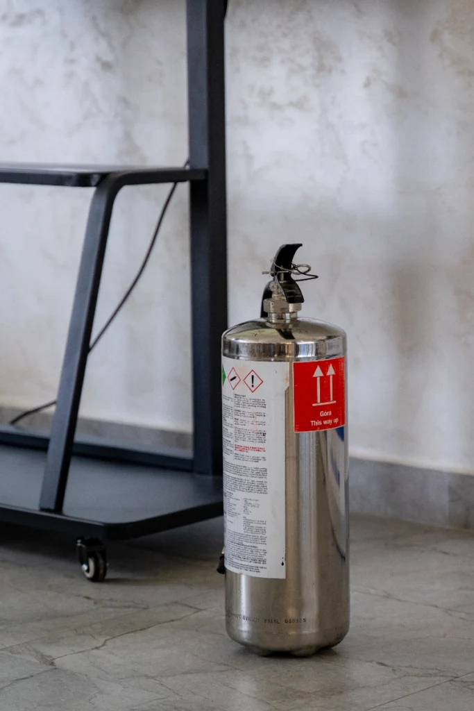 Silver fire extinguisher with a red “Góra This way up” sticker on the floor beside the screen stand