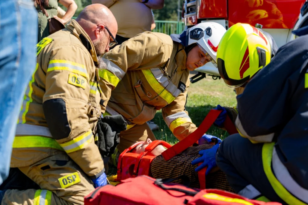Volunteer firefighters kneel on the grass by a casualty, fastening red straps and a head immobiliser