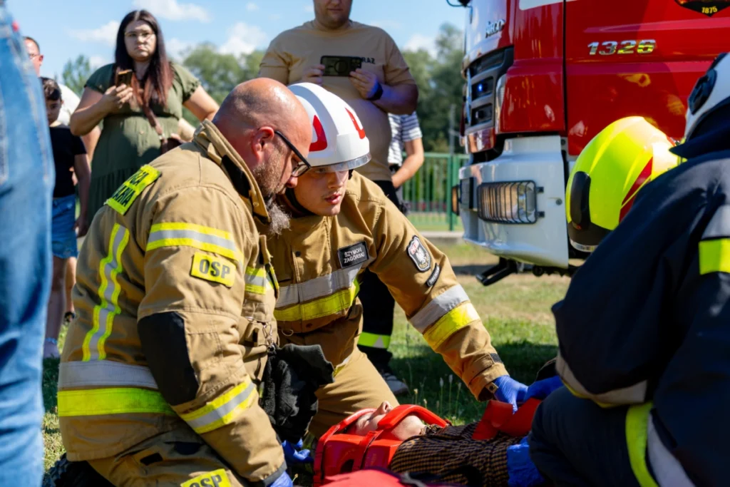 Volunteer firefighters bend over a casualty beside a fire engine as spectators film it on their phones