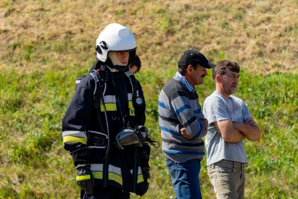 A white-helmeted firefighter with breathing apparatus waits on the grass beside two spectators with folded arms