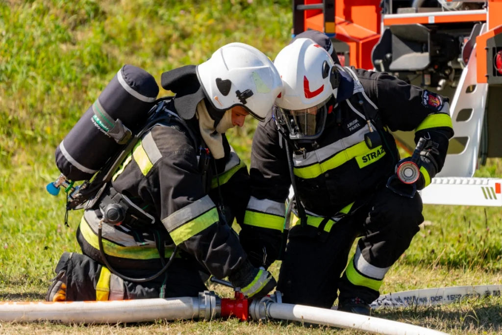 Two volunteer firefighters wearing breathing apparatus kneel on the grass and connect fire hoses