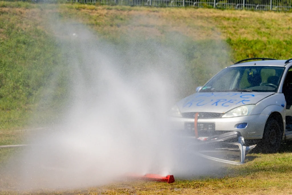 A red water curtain bar on the grass sprays in front of a silver car marked AUTO KUREK