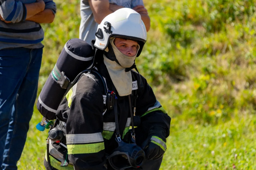 A white-helmeted firefighter with a breathing apparatus cylinder on his back and the mask hanging on his chest