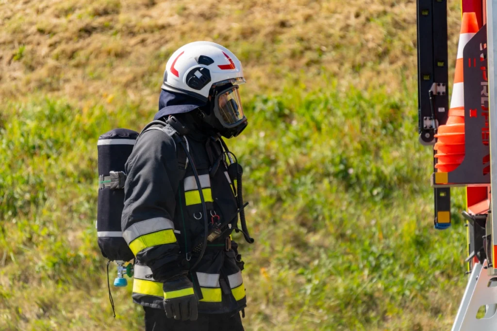 A firefighter in breathing apparatus, mask on, stands by a fire engine with orange traffic cones
